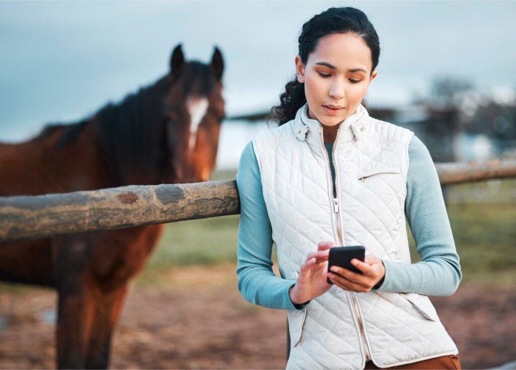 Woman standing with a bay horse while checking her phone.